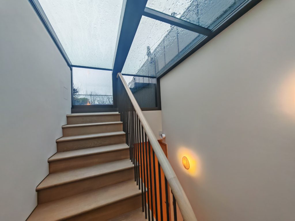 Wooden staircase with a black handrail leads up to a landing where sliding box rooflights and a large glass ceiling let in soft, overcast light through raindrops on the exterior window.