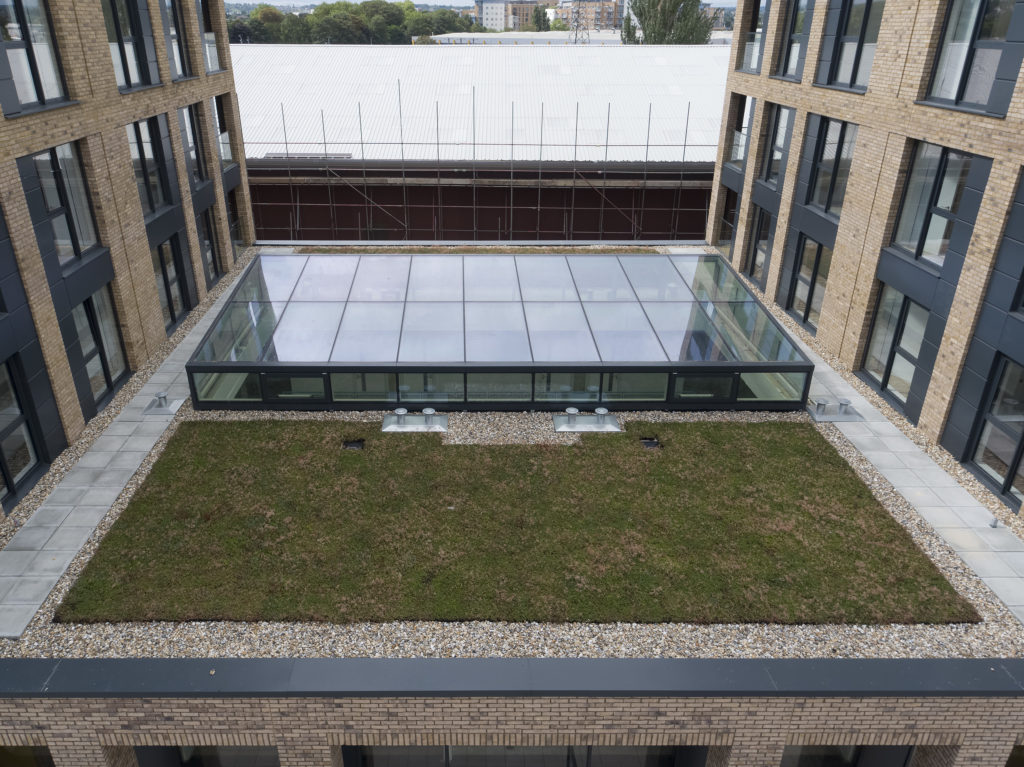 Rectangular courtyard with a green roof and glass skylight, featuring structural atria glazing, surrounded by brick buildings and paved pathways.