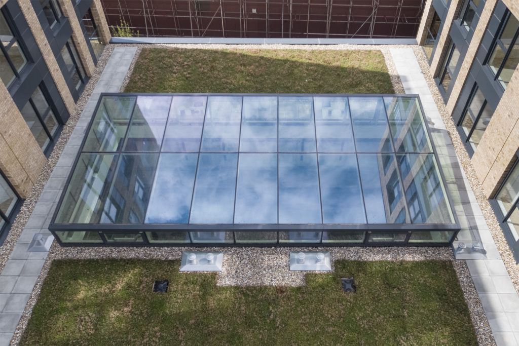 Aerial view of a rectangular structural atria glazing roof set in a grassy courtyard, surrounded by modern building walls with windows.