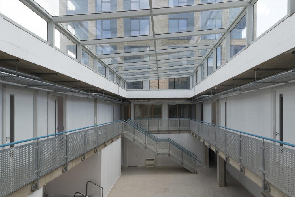 Interior view of a modern building featuring structural atria glazing, two floors, metal railings, and a central staircase connecting the upper and lower levels.