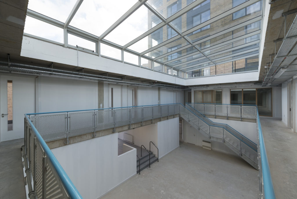 A modern building interior featuring structural atria glazing, a glass ceiling, metal railings, and a central staircase leading to an open upper level with white walls and unfinished floors.