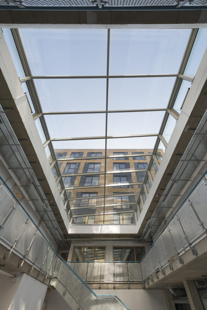 Looking up at a glass ceiling atrium with metal railings, the structural atria glazing frames a view of an adjacent brick building through the skylight.