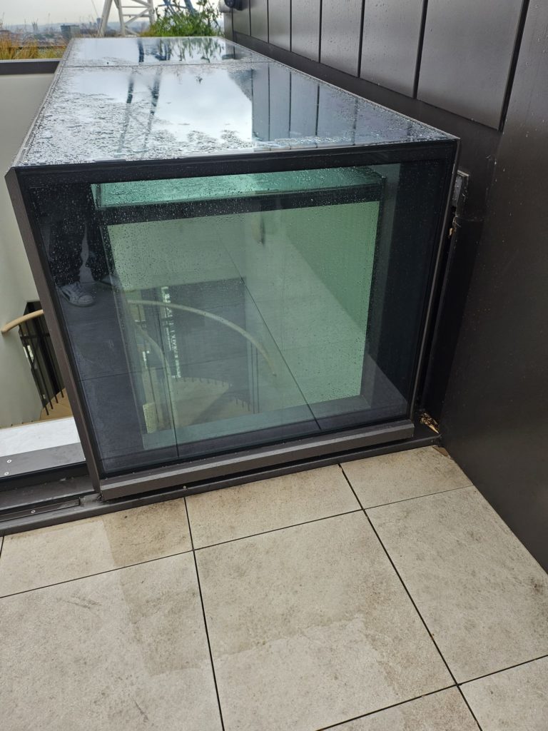 A glass enclosure with sliding box rooflights extends from a modern building's exterior, raindrops on its surface revealing a view of the spiral staircase inside.
