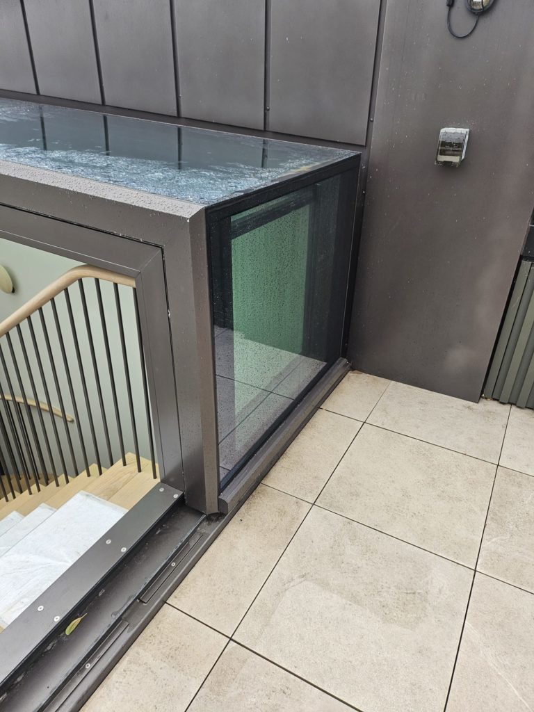 Glass-enclosed stairwell with a flat roof covered in rainwater, featuring sliding box rooflights, located next to a tiled outdoor area and a metal wall with a small light fitting.