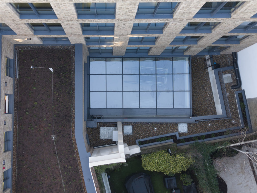 Aerial view of a modern building with bespoke commercial rooflights over a glass-covered atrium, adjacent rooftop areas with gravel, and a small green garden with shrubs and trees.