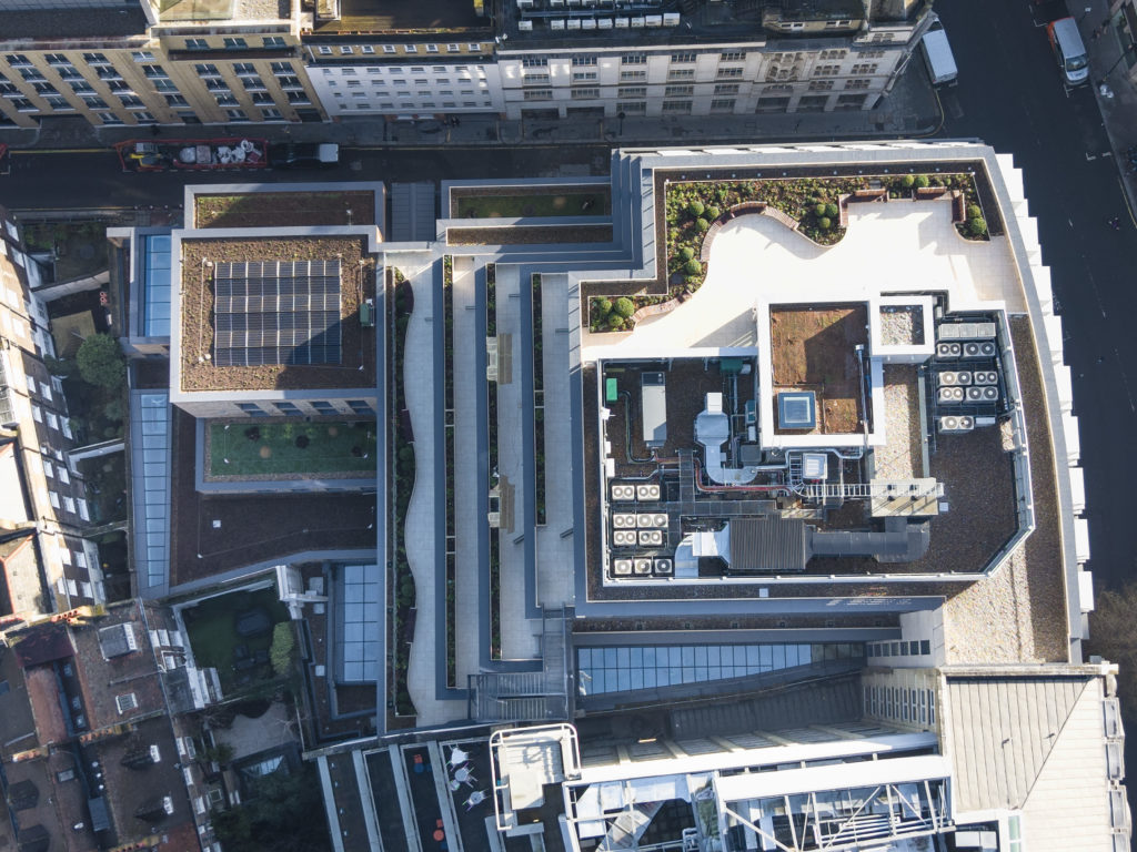 Aerial view of a modern city building complex with rooftops featuring gardens, solar panels, bespoke commercial rooflights, and ventilation systems, surrounded by adjacent streets and structures.