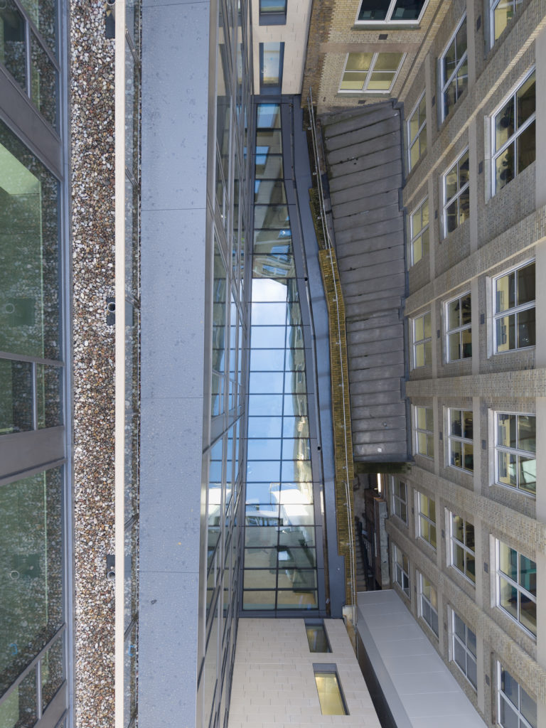 A glass skybridge connects two modern buildings with brick and stone exteriors, featuring bespoke commercial rooflights; windows reflect the sky above.