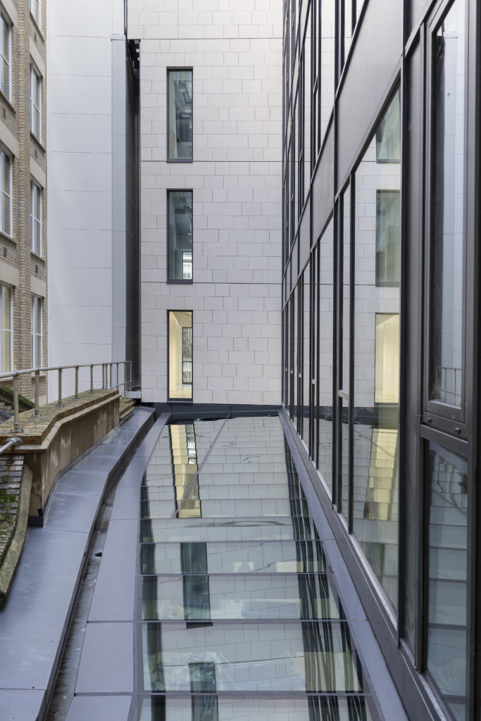 Modern building façade with large glass windows and bespoke commercial rooflights reflecting an adjacent structure; rectangular windows visible on the opposite wall.