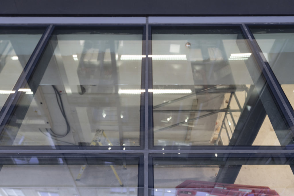 Large glass panels, designed as bespoke commercial rooflights, form a section of a floor, revealing an industrial interior below with a ladder, lights, pipes, and construction materials visible through the transparent surface.