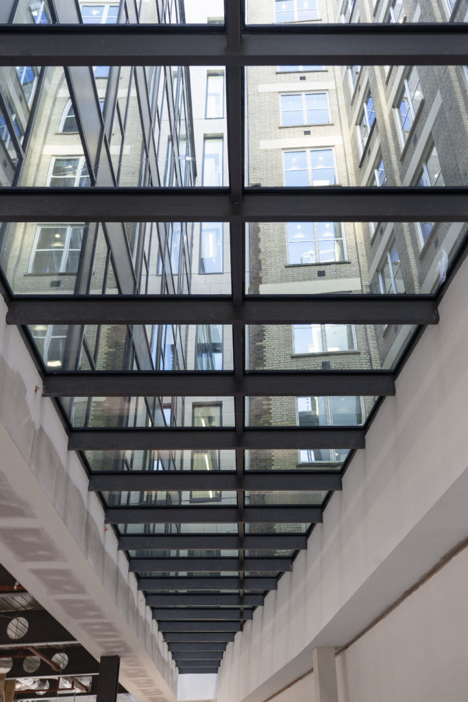 View looking up at a glass ceiling with metal beams, featuring bespoke commercial rooflights that reflect adjacent building windows and brick walls in an urban interior space.