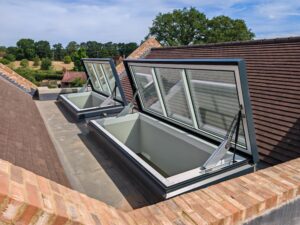 Two modern roof windows and a large access hatch are open on a sloping tiled roof, revealing built-in window wells with trees and fields visible in the background.