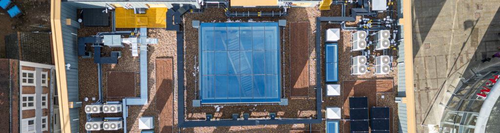 Aerial view of a building’s rooftop featuring a central glass structure with multipart fixed rooflights, multiple HVAC units, and various mechanical equipment.