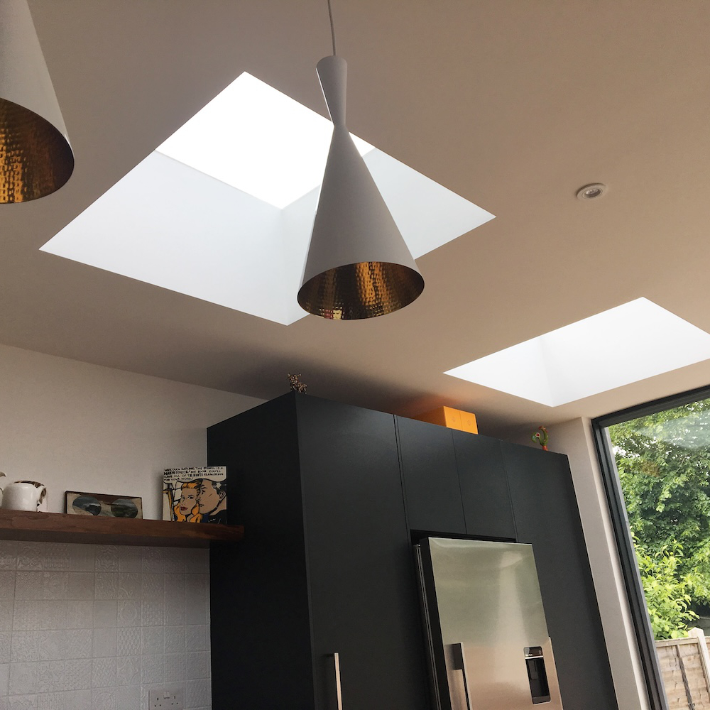 A kitchen with two skylight windows, cone-shaped pendant lights, dark cabinets, a stainless steel fridge, and a window with a view of greenery outside.