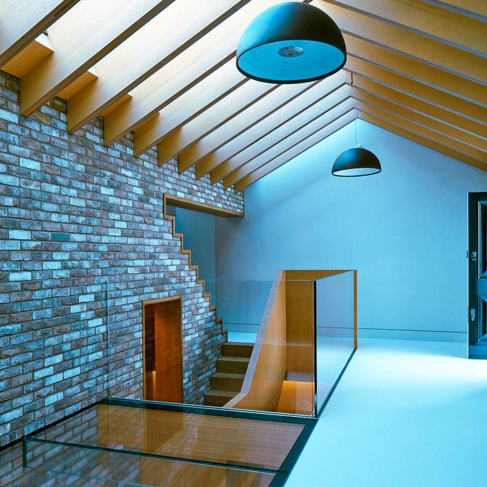 A modern interior with a glass railing staircase, exposed brick wall, wooden ceiling beams, and large pendant light fixtures under a slanted roof.