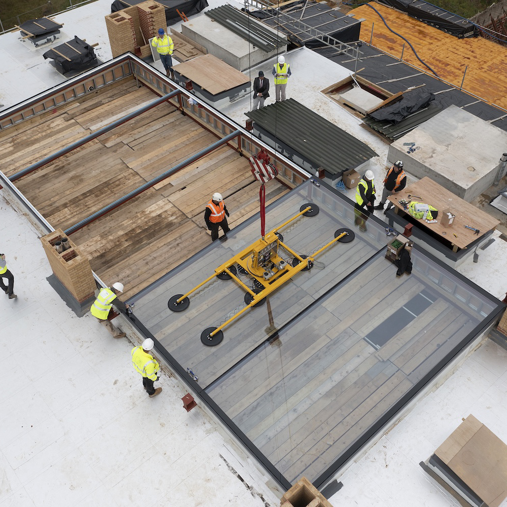 1000-IW-010921-AGi-136 Construction workers in safety gear install large glass panels on a building roof using a yellow lifting device. Scaffolding and building materials are visible around them.