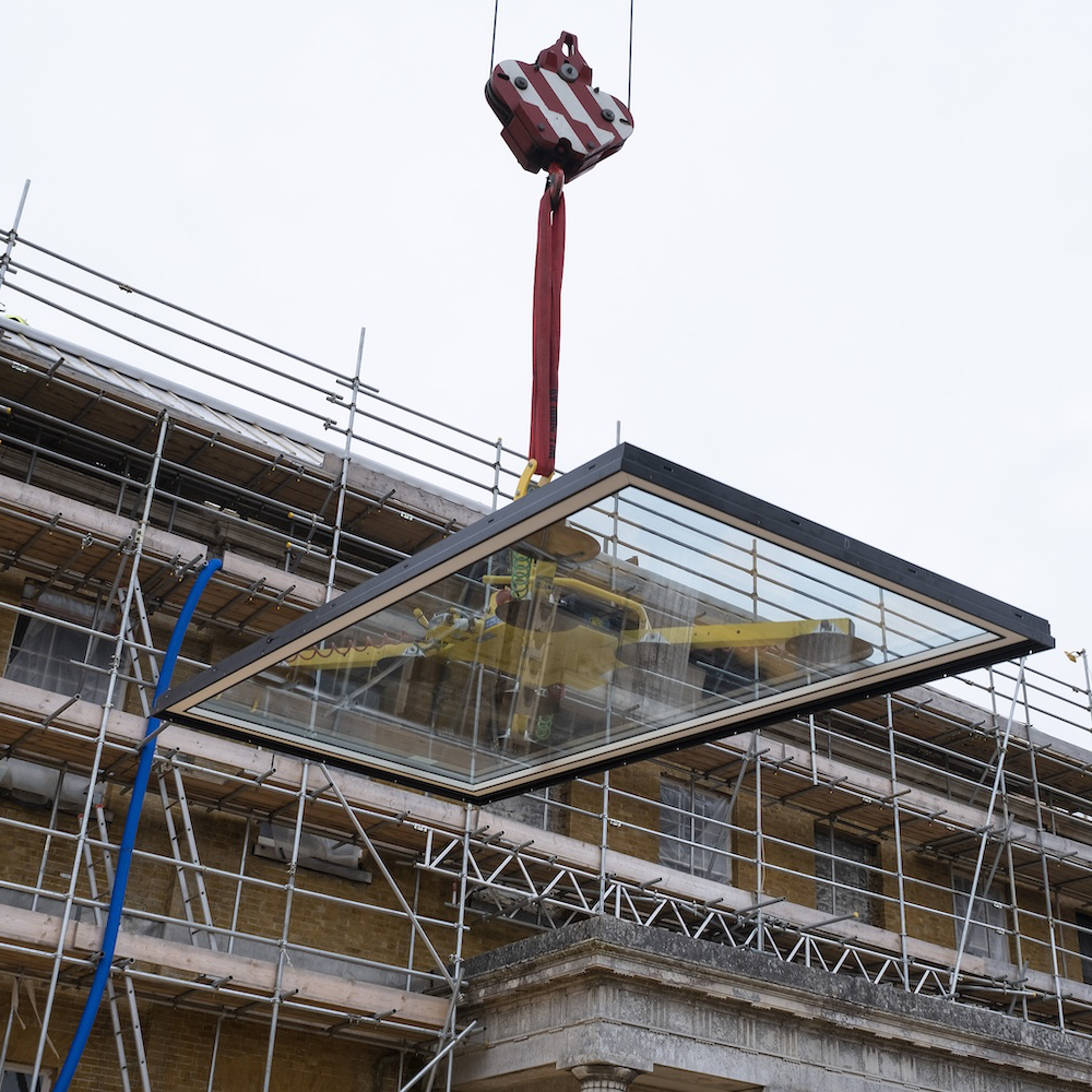 1000-IW-010921-AGi-145 A large glass panel is being hoisted by a crane in front of a building under construction, surrounded by scaffolding.