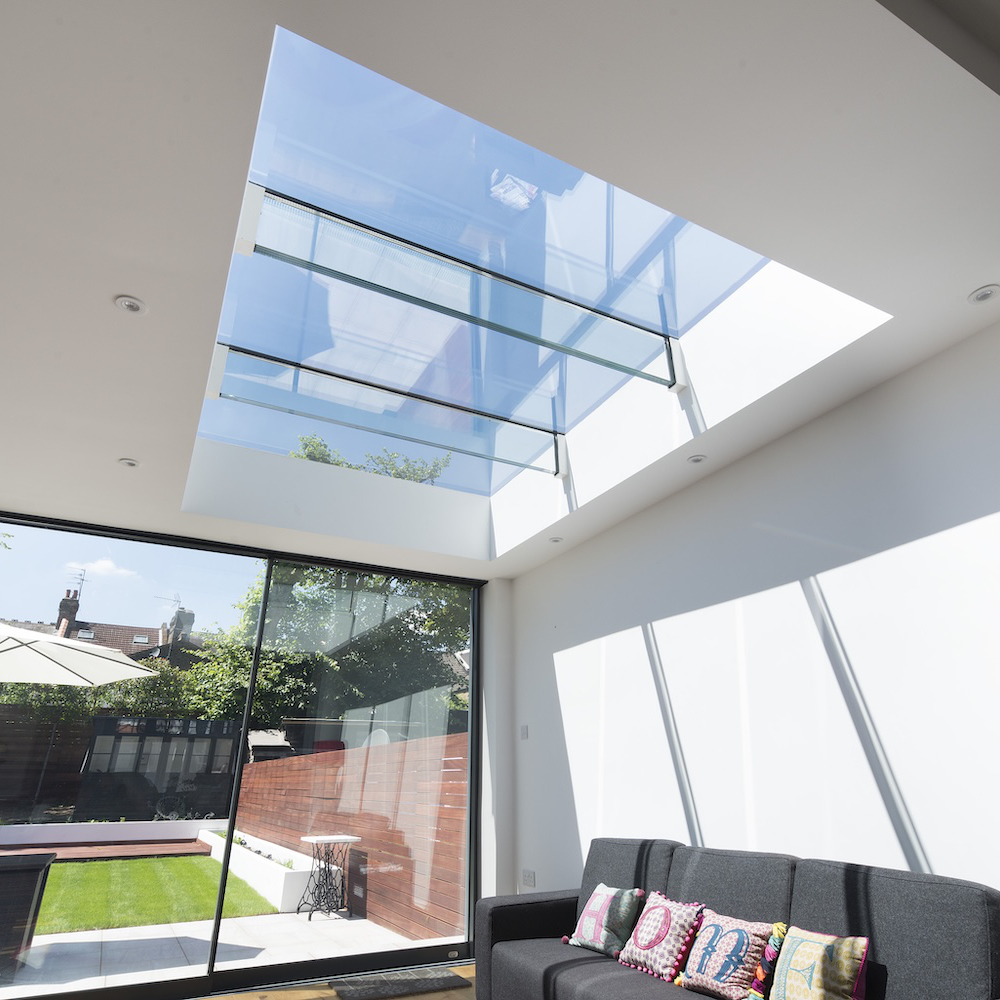 A modern room features a large skylight, a glass wall opening to a backyard with a pool, a patio, and a black sofa with colorful cushions.