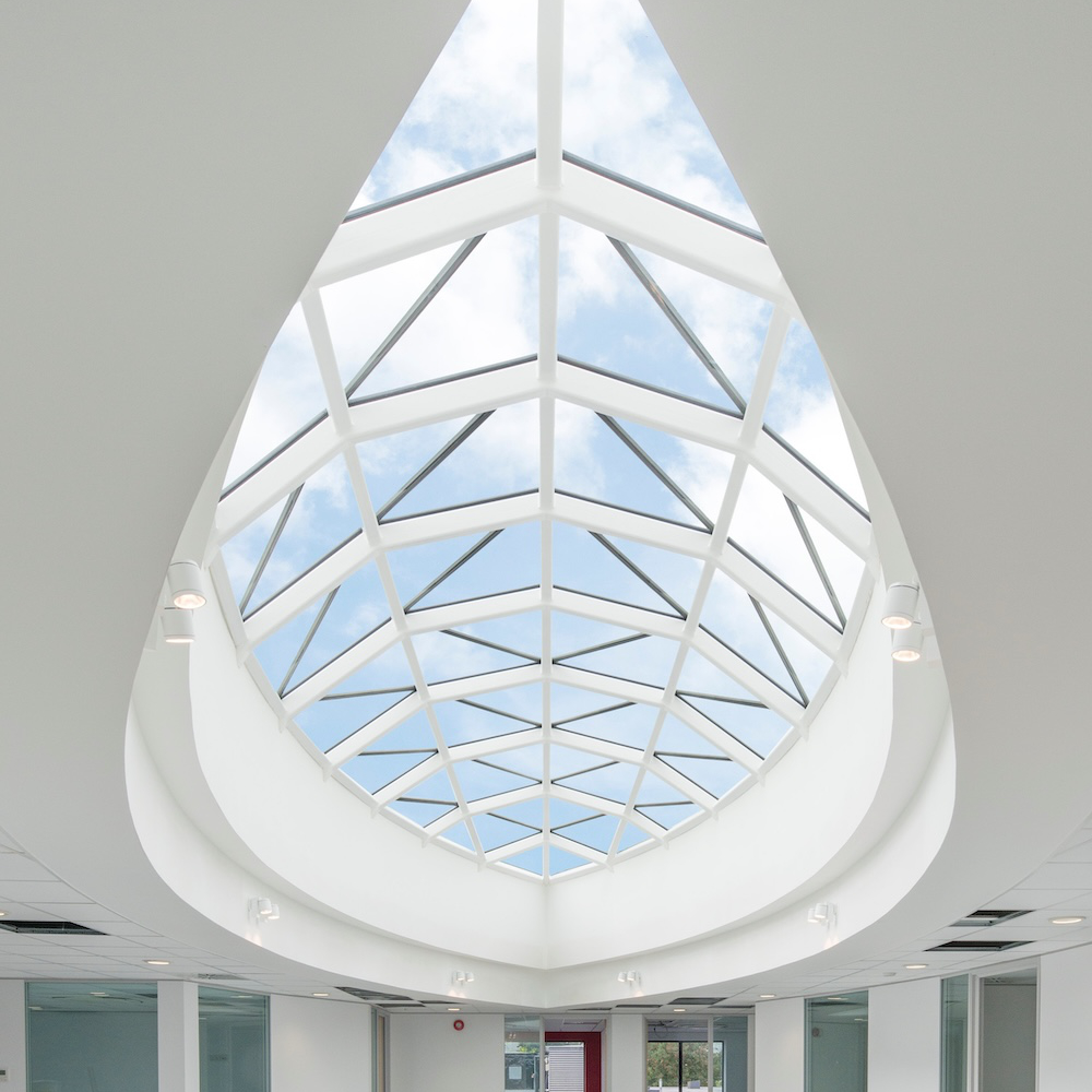 A contemporary interior space features a large, geometric glass skylight ceiling that reveals a view of the blue sky and clouds. The room has white walls and ceiling lights.