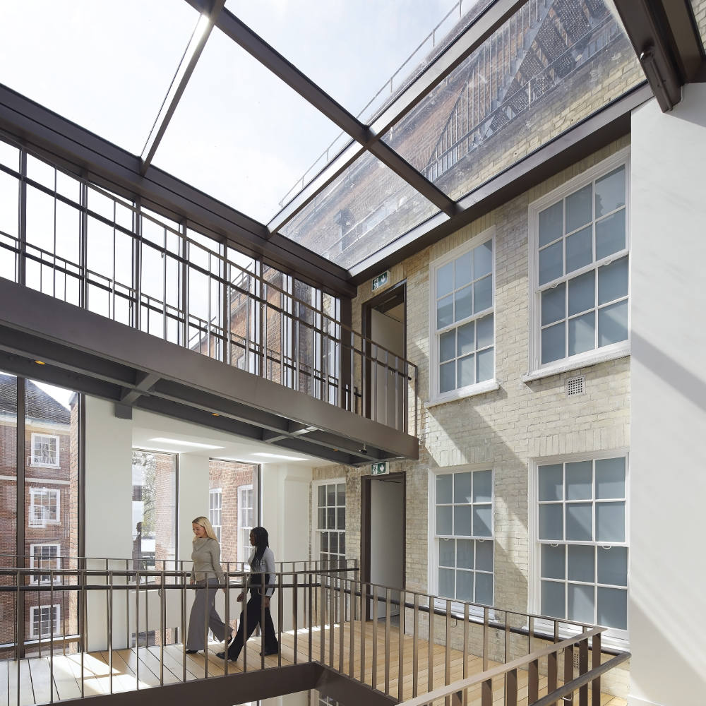 Two people walking along a walkway inside a modern building with large windows and exposed brick walls, under a glass ceiling, allowing natural light to illuminate the interior space.