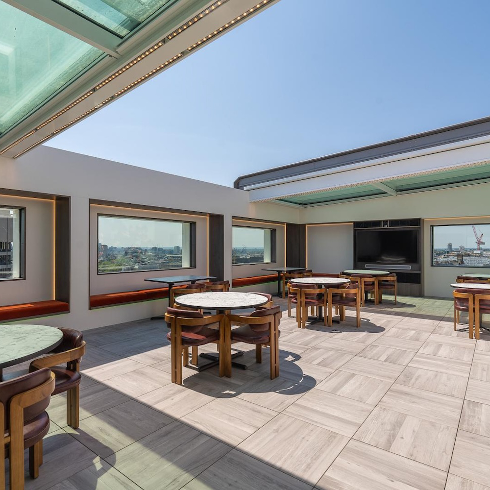 A rooftop seating area with round tables and chairs, enclosed by large windows and a retractable glass roof on a sunny day.