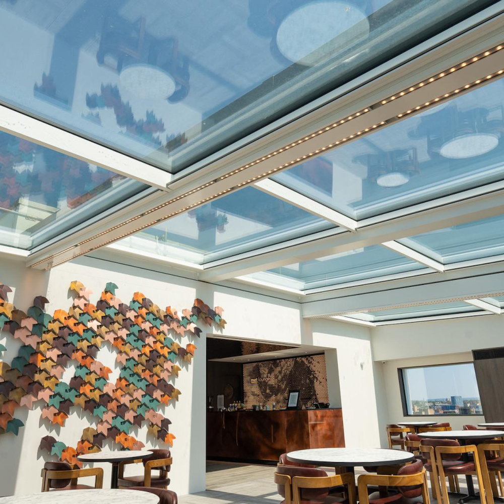 A modern indoor seating area with glass ceilings, circular wooden tables, chairs, and a colorful wall art installation consisting of leaf-shaped decorations.