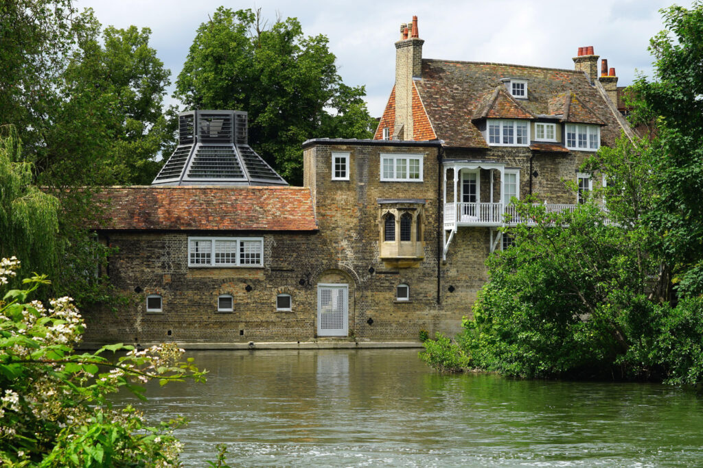 Historic brick building with a tall chimney and a glass-roofed structure on the left, situated alongside a tranquil river and surrounded by lush greenery.
