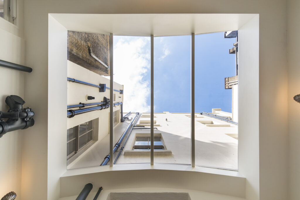 View of the sky through vertical skylights in an indoor courtyard surrounded by tall buildings. Pipes and structural elements are visible on the walls.