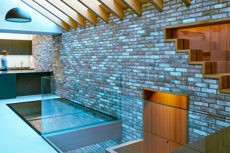 Modern interior with exposed brick wall, glass floor panels, and a person near a kitchen area in the background. The ceiling features wooden beams and a skylight.