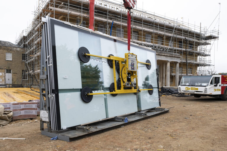 A crane lifts a large glass panel in front of a building under construction, surrounded by scaffolding and construction materials.