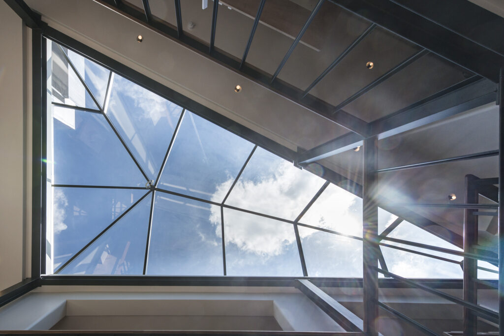 View of the sky through a geometric, glass ceiling from inside a modern building, with metal framework and a few light fixtures.