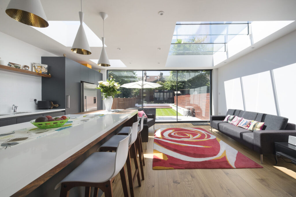 Modern kitchen and living area with large windows, white countertops, dark cabinetry, a decorative rug, and glass doors leading to an outdoor patio with greenery.
