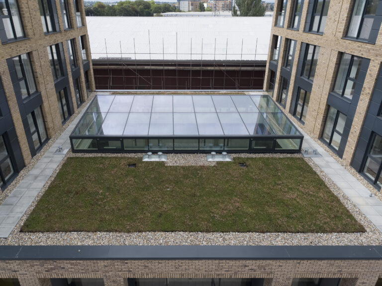 A view of a rectangular courtyard between buildings, featuring a central area with grass and a glass-roofed section. The surrounding buildings have brick exteriors and multiple windows.