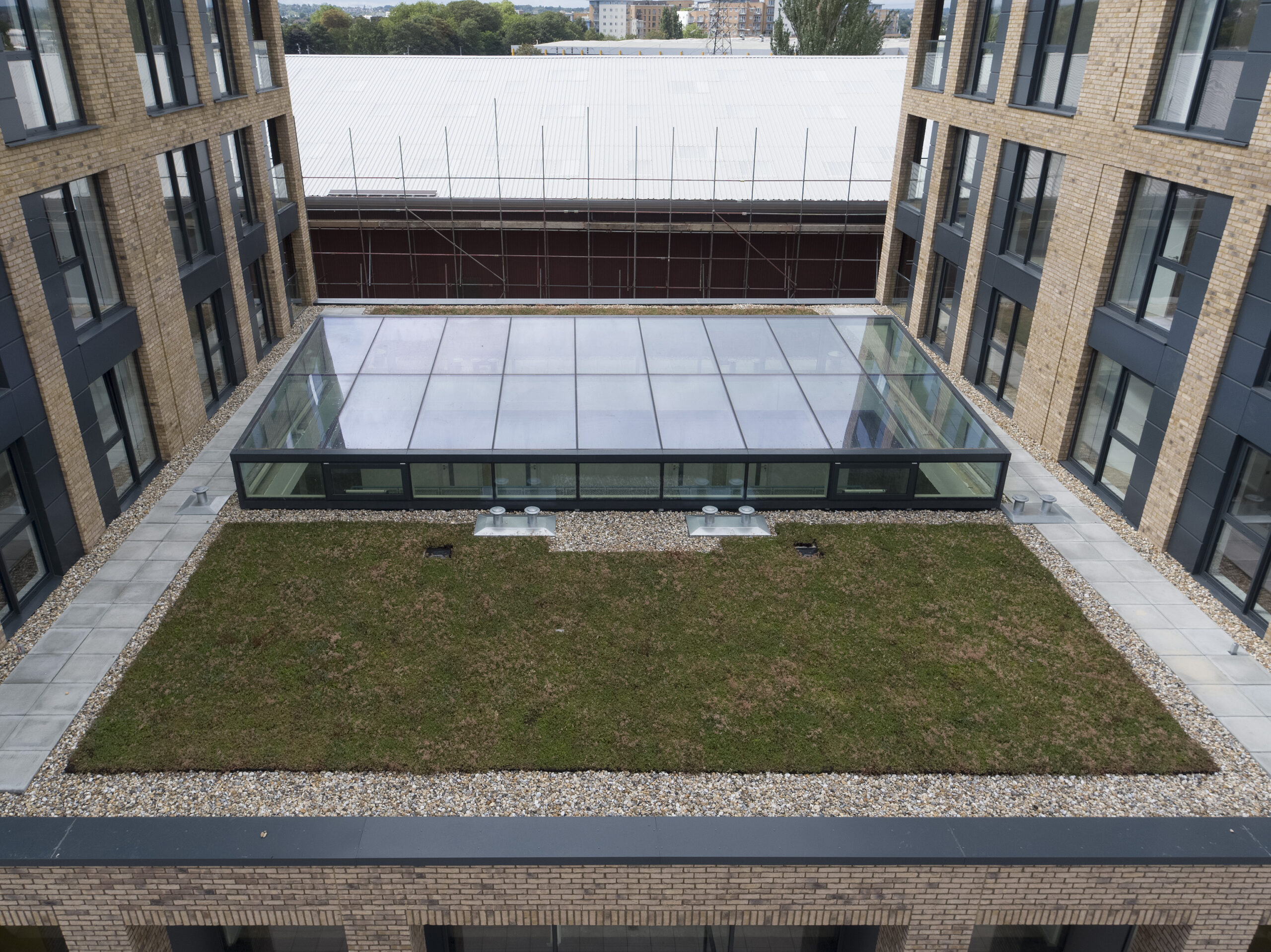 A view of a rectangular courtyard between buildings, featuring a central area with grass and a glass-roofed section. The surrounding buildings have brick exteriors and multiple windows.
