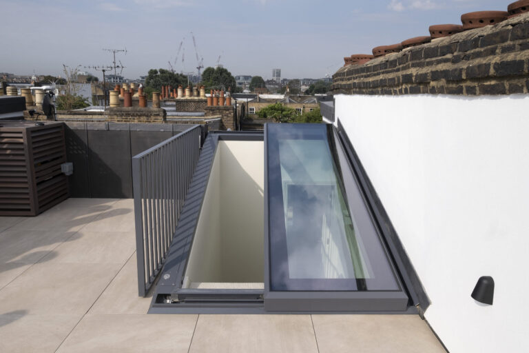 A rooftop with a large glass skylight and a metal railing, leading to an interior staircase. Brick chimneys and cityscape are visible in the background.