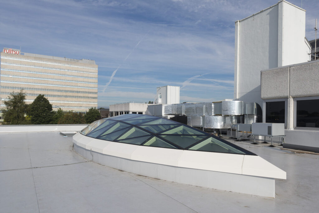A rooftop with a modern glass skylight structure, adjacent to ventilation systems and neighboring buildings, including a visible Fujitsu building in the background under a blue sky with wispy clouds.