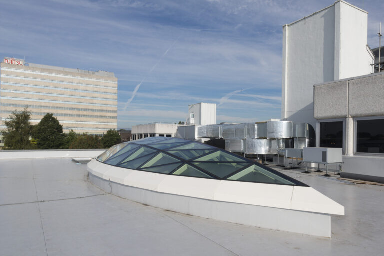A rooftop with a modern glass skylight structure, adjacent to ventilation systems and neighboring buildings, including a visible Fujitsu building in the background under a blue sky with wispy clouds.