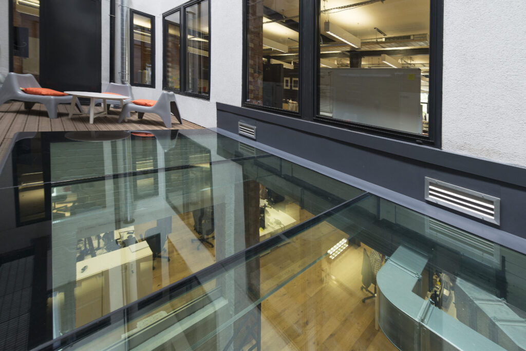 Office building with a glass floor, revealing an interior workspace below. The space is furnished with chairs and desks, and an outdoor area with patio chairs and a table is also visible.
