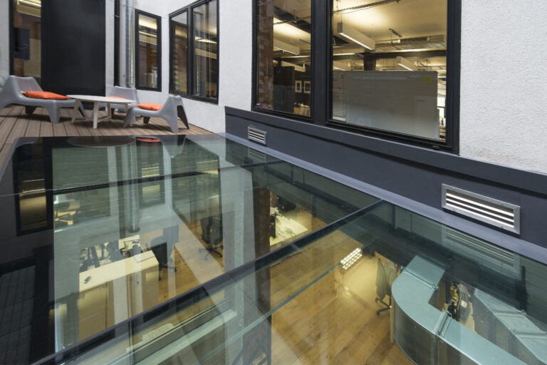 Office building with a glass floor, revealing an interior workspace below. The space is furnished with chairs and desks, and an outdoor area with patio chairs and a table is also visible.