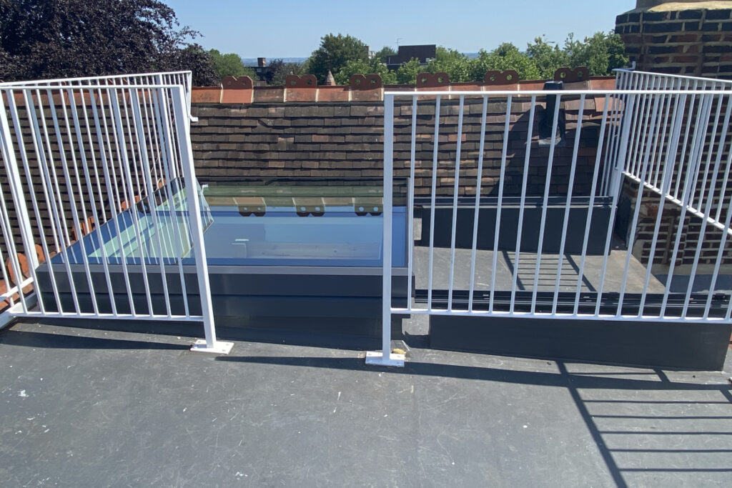 A rooftop with a brick wall, metal railings, and a glass skylight enclosure, viewed under a clear sky.