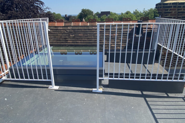 A rooftop with a brick wall, metal railings, and a glass skylight enclosure, viewed under a clear sky.