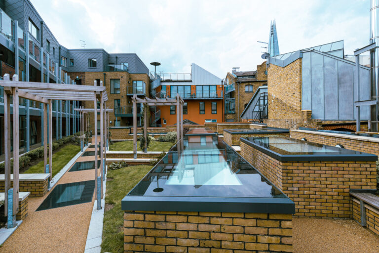Modern courtyard with brick buildings, glass structures, and water features. Pathways and seating areas are visible. The Shard skyscraper can be seen in the background.
