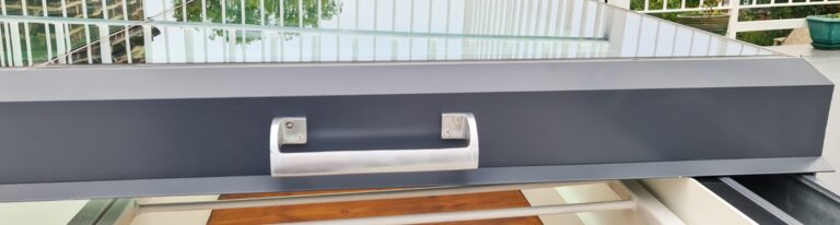 A close-up view of a metal handle on a railing structure with glass panels and greenery in the background.