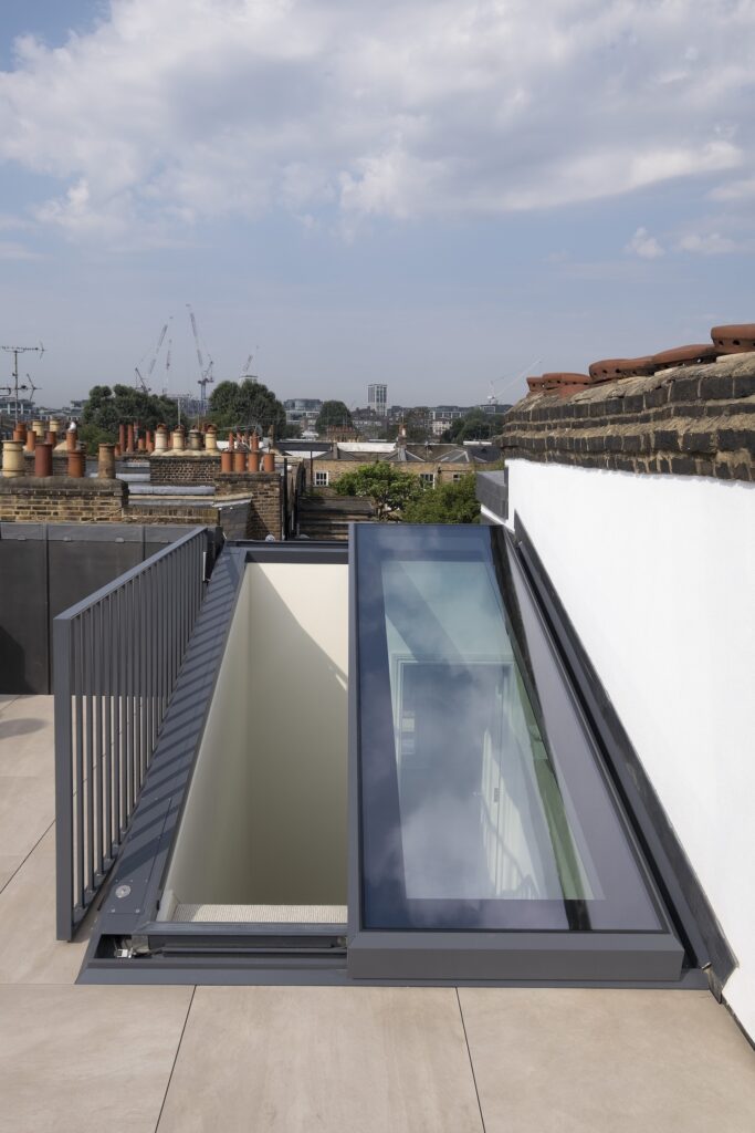 A rooftop terrace with a large glass skylight and metal railing, overlooking a cityscape with buildings and cranes in the distance under a partly cloudy sky.
