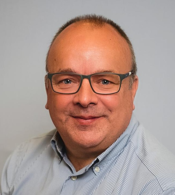Alan Saxby with glasses, short hair, and a slight smile, wearing a light-striped button-up shirt, poses for a headshot against a plain background.