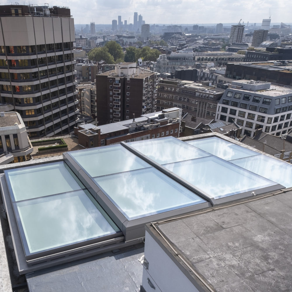 A rooftop access hatch with a glass cover and safety railing, surrounded by fencing, on a building's flat roof.