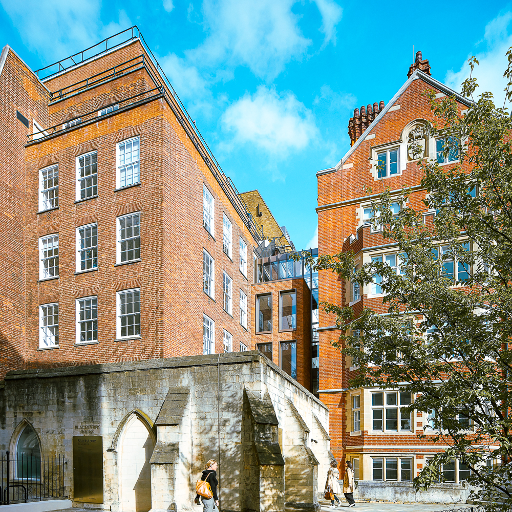 Two large brick buildings under a blue sky, with an arched stone structure and people walking in the foreground.
