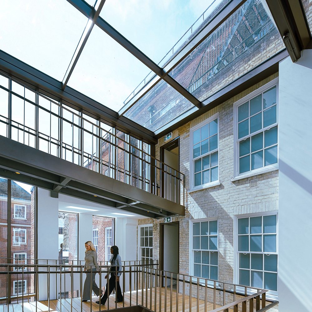 Two people walk across a glass-covered skybridge in a building with brick walls and large windows.