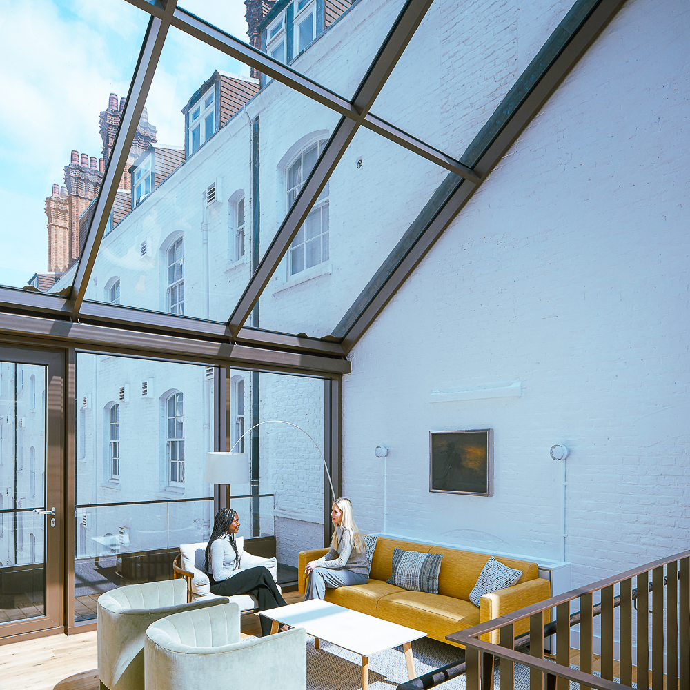 Two people sit on a sofa in a bright room with a glass ceiling, surrounded by modern furniture and white brick walls.