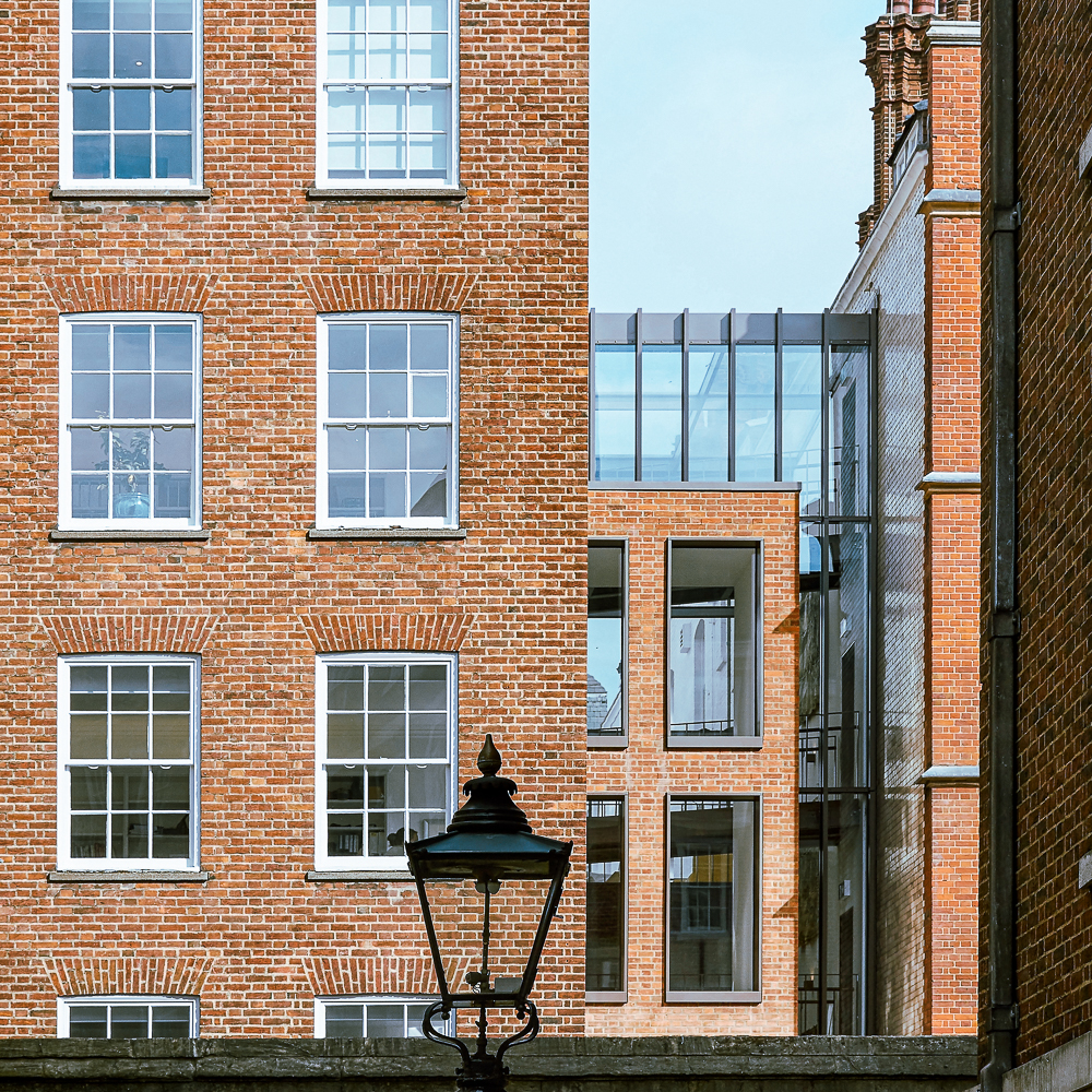 A view of two adjacent red brick buildings with multiple windows, separated by a modern glass corridor. A vintage street lamp is visible in the foreground.