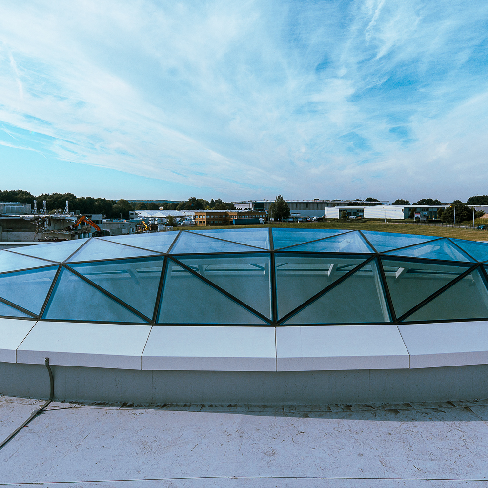 A geometric glass roof with triangular panels, set against a backdrop of a blue sky and distant buildings, viewed from a rooftop.